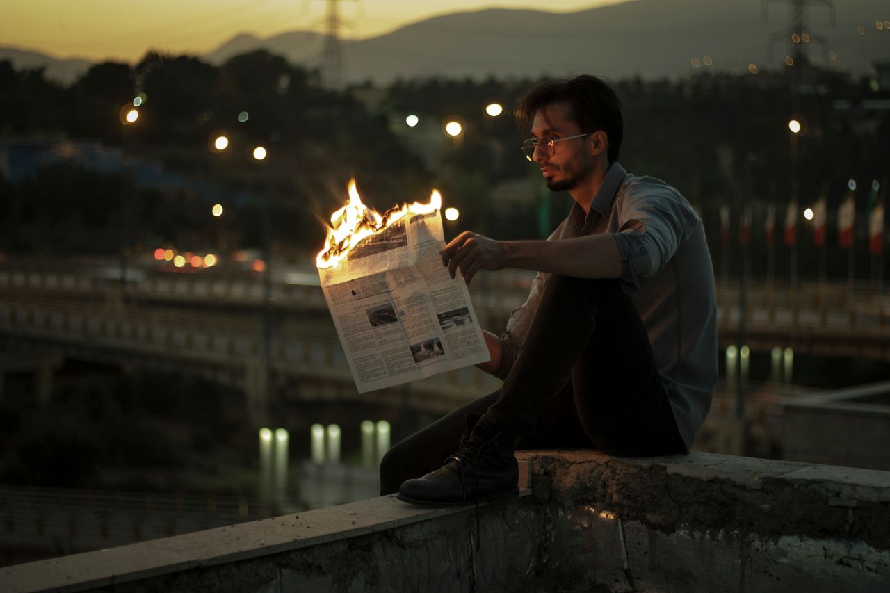 A man sitting outdoors at sunset, holding a burning newspaper with an urban backdrop.