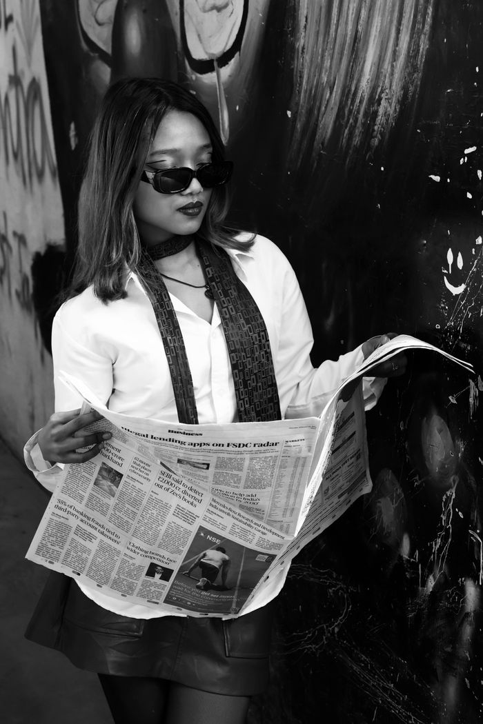 Black and white photo of a fashionable woman in sunglasses reading a newspaper in New Delhi.