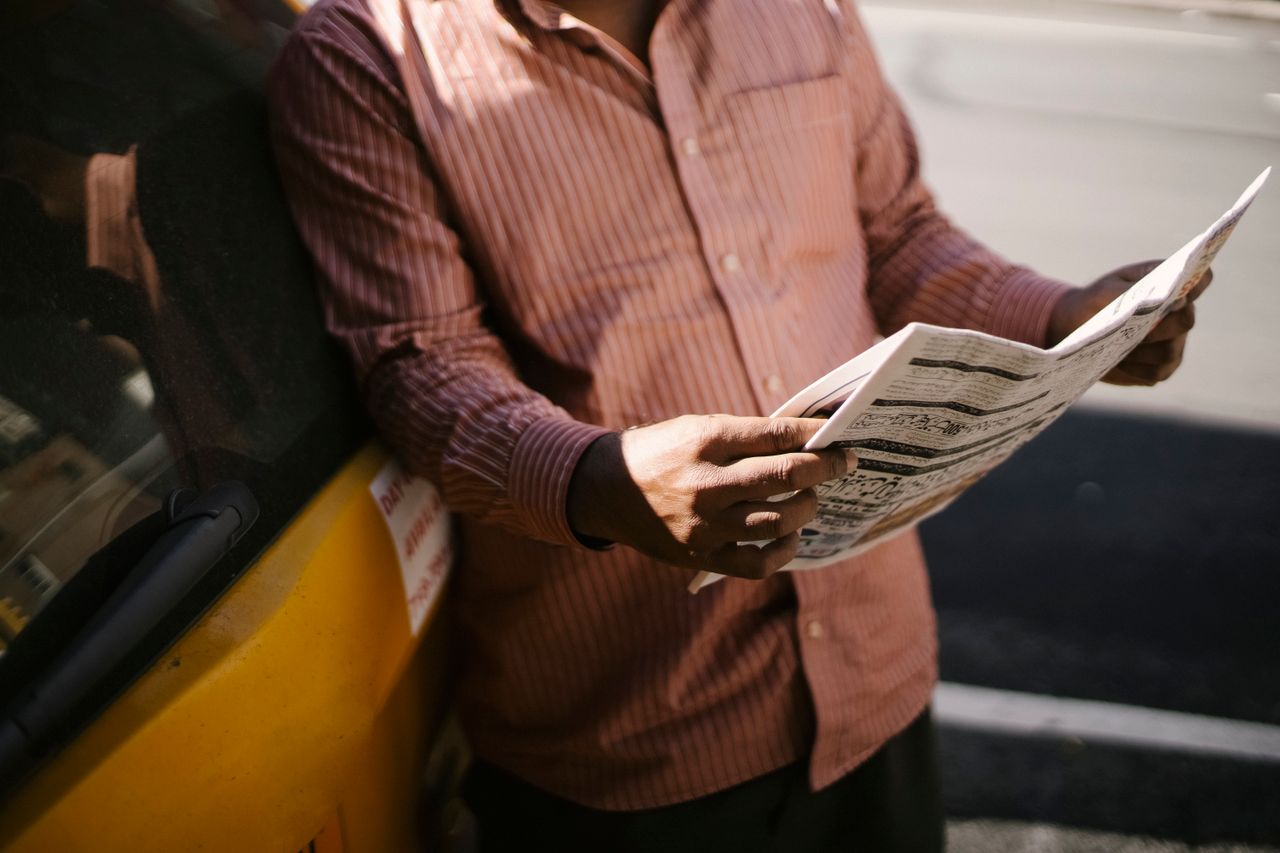 Crop anonymous ethnic cab driver in ornamental shirt standing with newspaper near transport in sunlight
